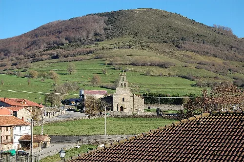 Iglesia de San Miguel de Aguayo en un paisaje rural con colinas y casas.