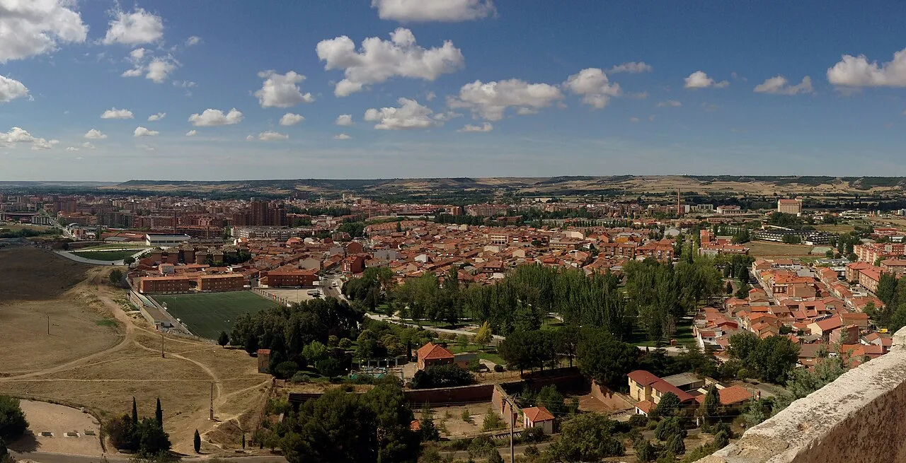 Panoramic view of Palencia with buildings and green areas under a cloudy sky.