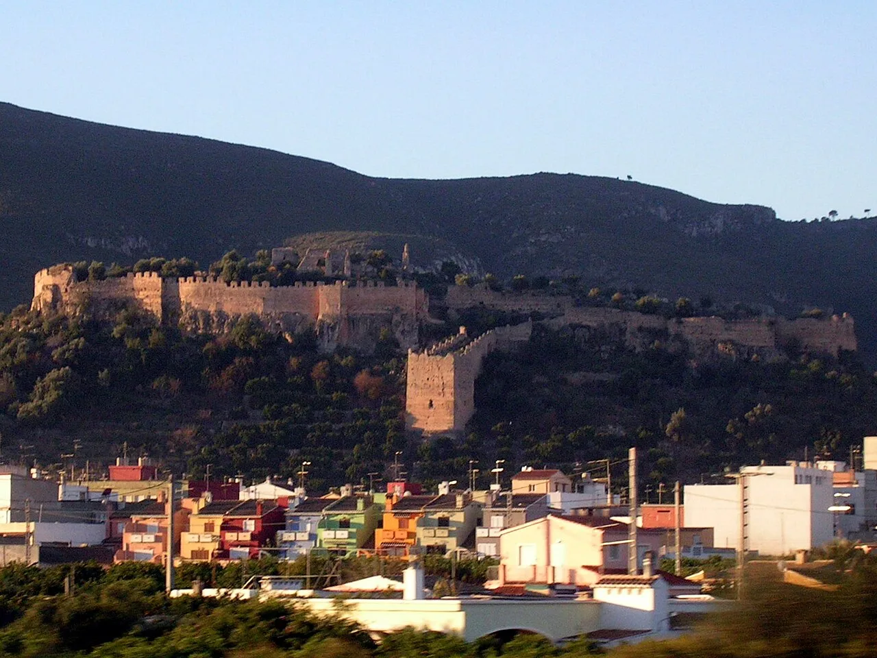 Castillo en lo alto de una colina con casas en primer plano y montañas al fondo.