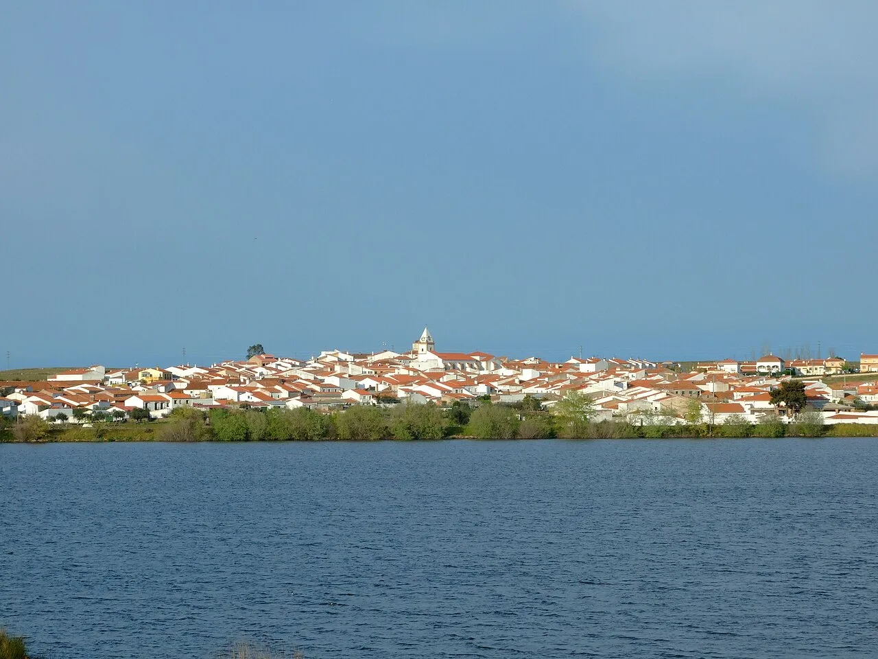 Blick auf Membrío vom Stausee, mit Häusern und einer Kirche im Hintergrund.