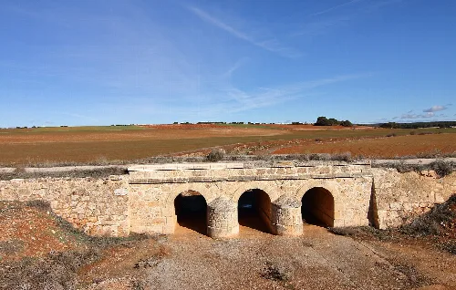 Steinbrücke mit drei Bögen in einer Landschaft aus Feldern und blauem Himmel.