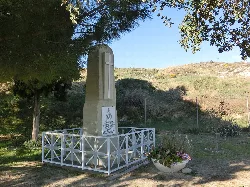 Denkmal mit Kreuz und Tafel in einem offenen Raum mit Bäumen und Gras.