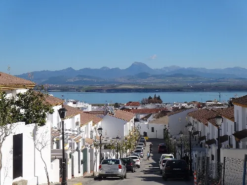 Straßenansicht in Bornos mit weißen Häusern und geparkten Autos, Stausee und Berge im Hintergrund.