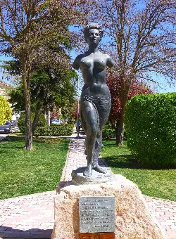 Skulptur einer nackten Frau in einem Park mit Bäumen und Sträuchern.