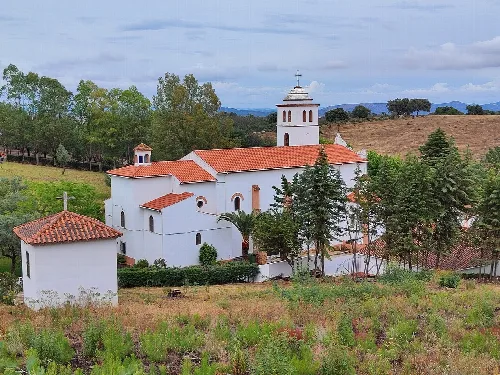 Weiße Kirche mit rotem Dach und Turm mit Kreuz in einer ländlichen Landschaft.
