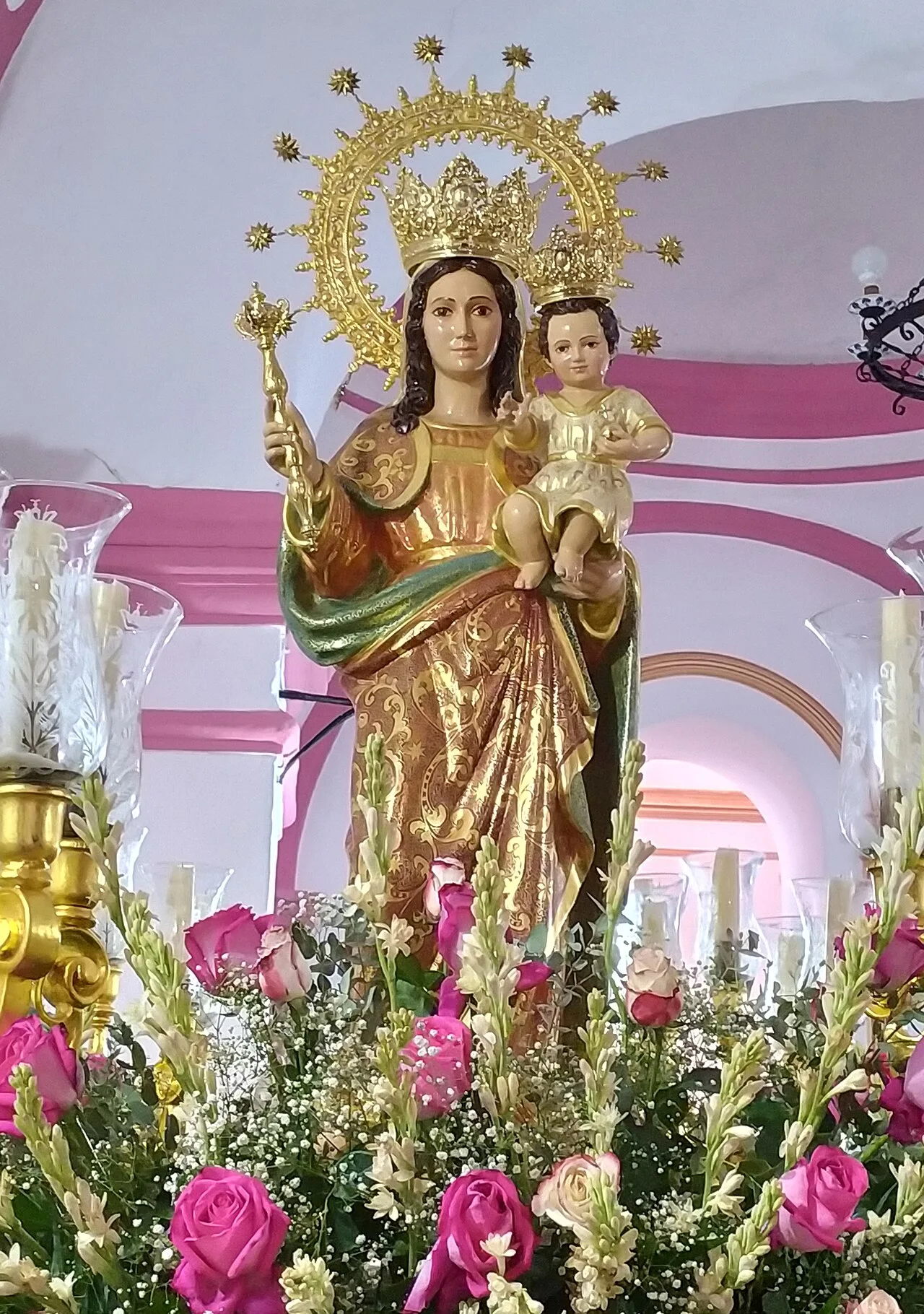 Estatua de la Virgen de los Remedios con un niño, rodeada de flores y velas.