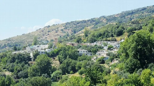 Landschaft mit weißen Häusern an einem Hang, umgeben von Bäumen und Bergen.