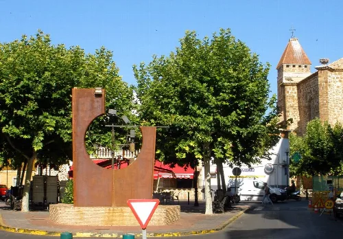 Escultura de metal en una plaza con árboles y una iglesia en el fondo.
