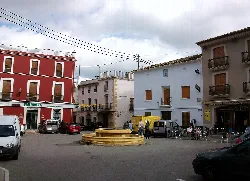 Square with central fountain, white and red buildings, and people and cars.