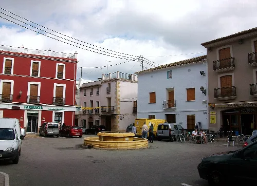 Platz mit zentralem Brunnen, weißen und roten Gebäuden, Menschen und Autos.