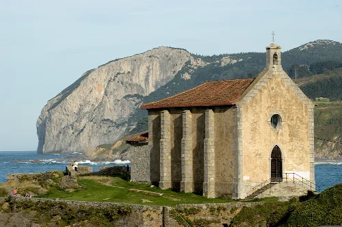 Pequeña iglesia de piedra junto al mar con acantilados en el fondo.
