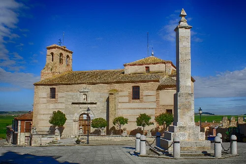 Iglesia de piedra con torre y monolito en la plaza del pueblo.
