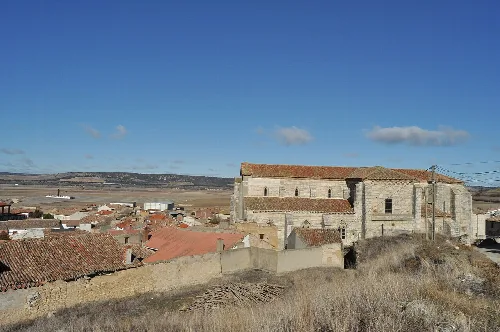 Church and traditional houses of Herrera de Valdecañas with countryside and blue sky in the background.