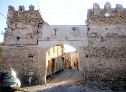 Arco de piedra con restos de muralla y una calle pequeña detrás.