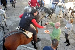 Person on a horse offering potatoes and wine in a square with people and horses.