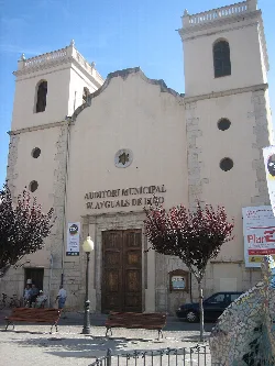 White building facade with towers and wooden doors, in a square with trees and benches.