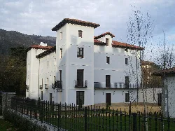 White building with multiple floors and towers, surrounded by a fence and gardens.