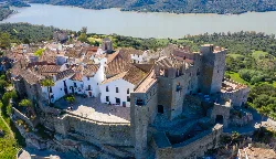 Vista aérea de un castillo medieval en una colina con casas blancas y un embalse en el fondo.