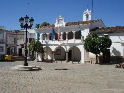 Square with white building, flags, trees, and lamppost in Aceuchal.