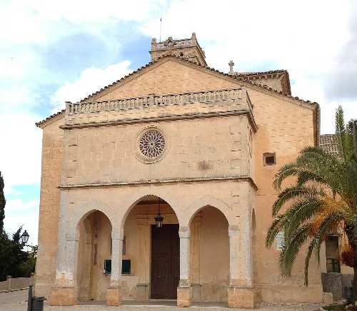 Steinfassade einer Kirche mit Bögen und einer Palme rechts in Ariany.