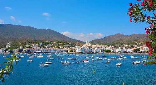 Hafen von Cadaqués mit Booten und weißen Häusern, Berge im Hintergrund.