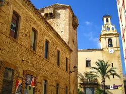 Tower and church in El Verger, with a clock on the tower and palm trees in the foreground.