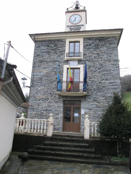 Edificio de piedra con reloj y banderas en Villanueva de Oscos.