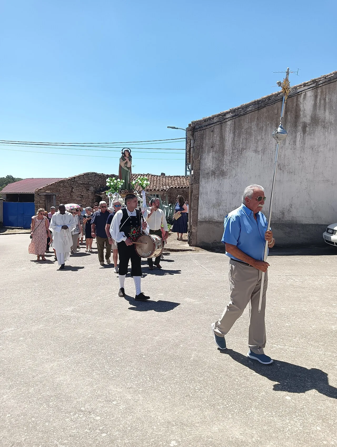 Hombre con bastón y cruz lidera una procesión con personas vestidas de fiesta en Barceo.
