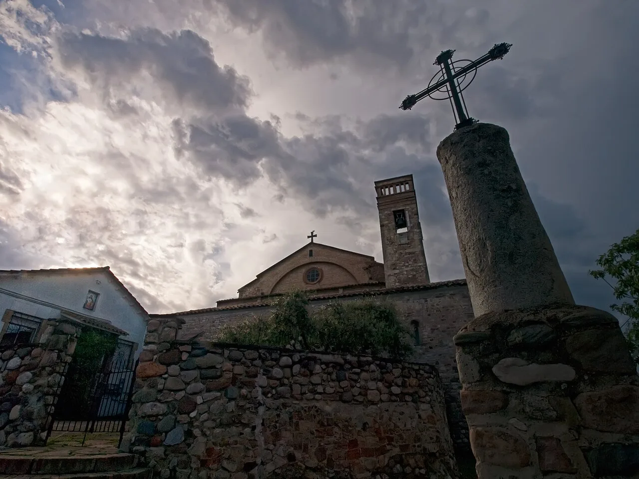 Kirche von Polinyà und ein Kreuz im Vordergrund mit bewölktem Himmel.