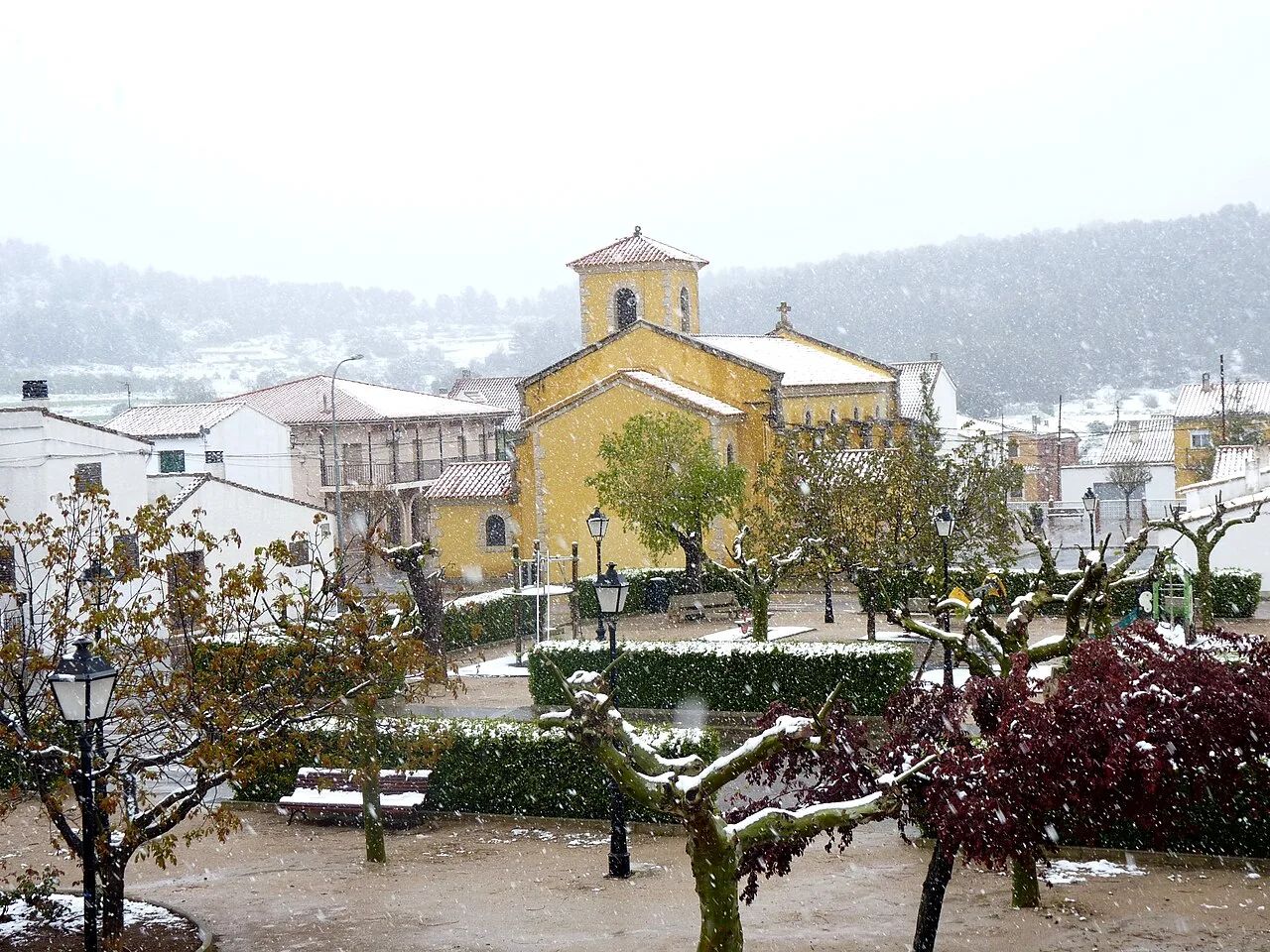 Iglesia y parque con árboles y bancos cubiertos de nieve en Benagéber.