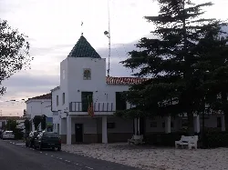 Edificio blanco con torre y bandera, rodeado de árboles y bancos en Marines.