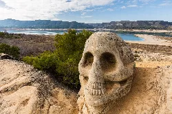 Schädelskulptur auf der Ruta de las Caras mit Stausee und Bergen.