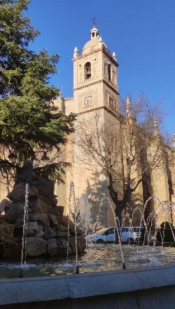 Torre de iglesia con reloj y campanario en Don Benito, con fuente y árboles en primer plano.