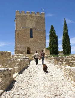 Tres personas y un perro caminando por un camino de piedra hacia una torre medieval.