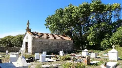Pequeña capilla de piedra con cruz en su tejado, rodeada de tumbas y árboles.