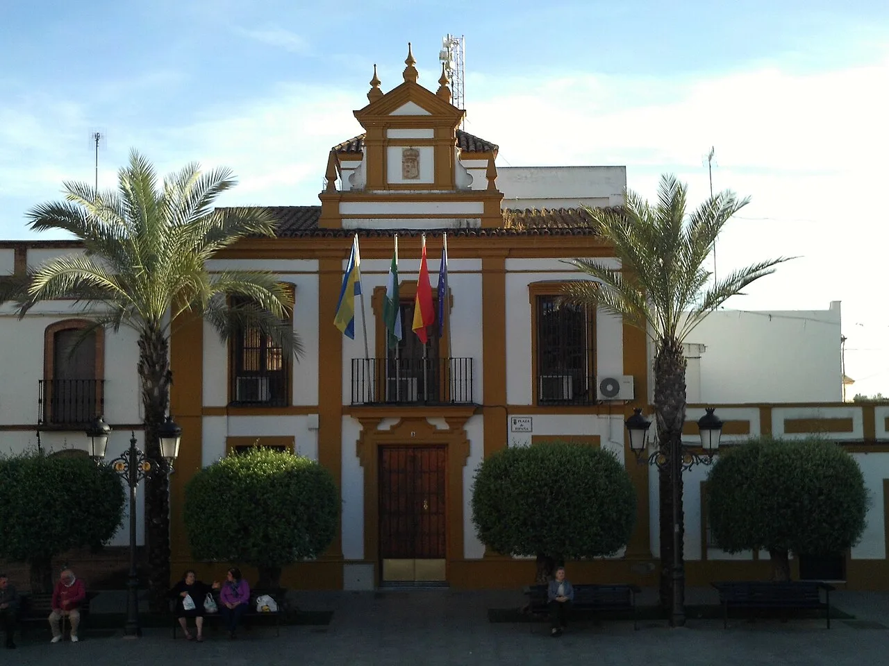 Edificio antiguo con banderas, palmeras y personas sentadas en la plaza.