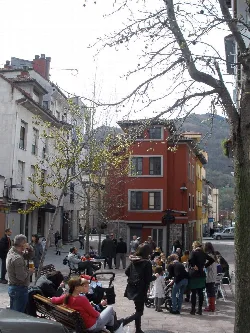 People sitting and standing in a square with trees and colorful buildings in Pola de Laviana.